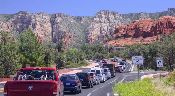 A long line of vehicles, including trucks, SUVs, and cars, is seen in traffic on a scenic highway surrounded by stunning red rock formations and towering cliffs. The clear blue sky and lush greenery contrast with the rugged desert landscape, capturing a classic road trip or tourist destination scene.