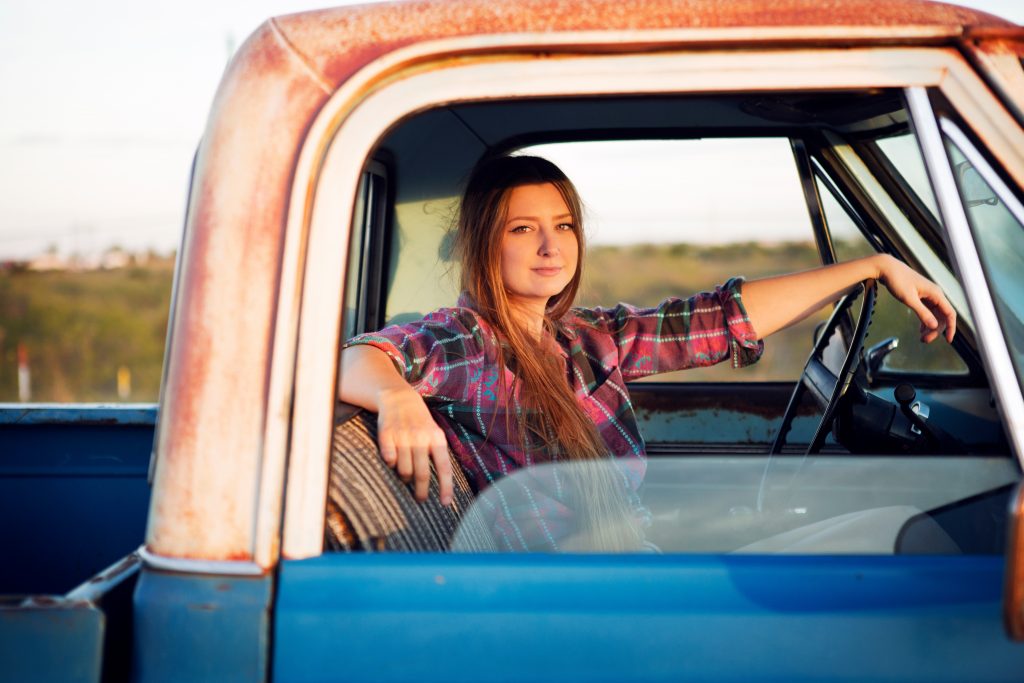 A young woman with long blonde hair, wearing a plaid shirt, sits confidently in the driver's seat of an old blue pickup truck with a rusted frame, parked in a rural setting during sunset.
