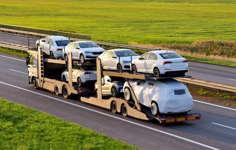 A large car carrier truck transporting multiple new vehicles on a highway, with green fields in the background. One of the vehicles is covered with a protective wrap.