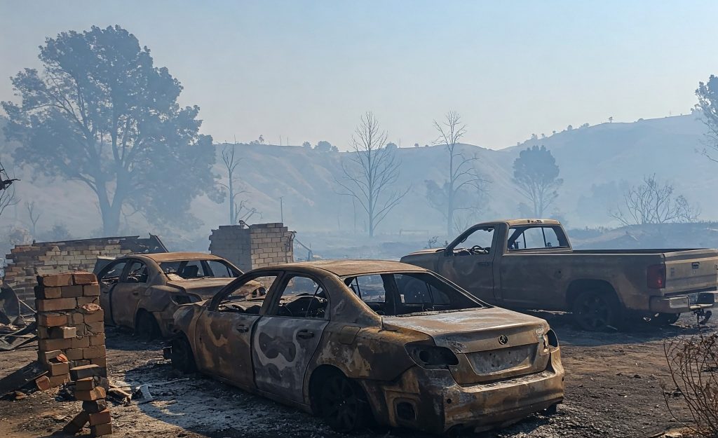 Burned-out vehicles, including a sedan and a pickup truck, in a charred landscape after a wildfire, with smoke lingering in the air and scorched hills in the background.