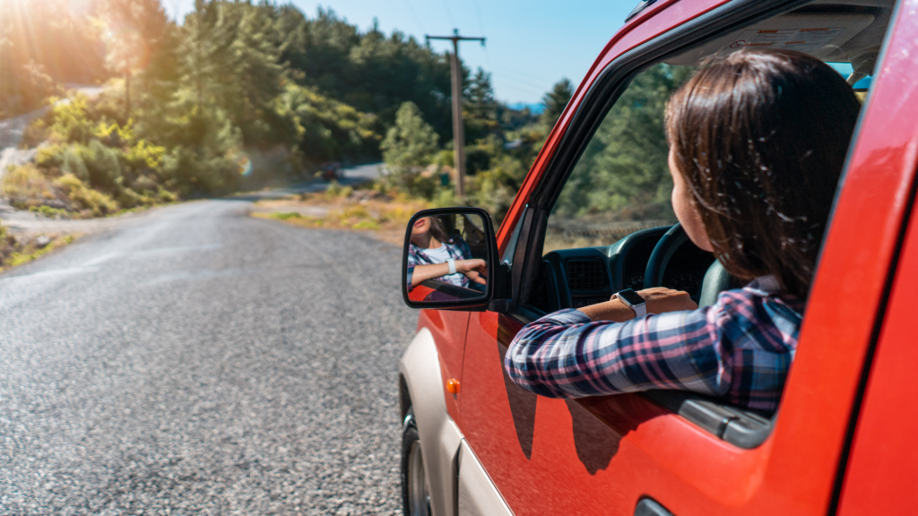 A woman with long dark hair, wearing a plaid shirt, drives a red SUV down a scenic rural road surrounded by lush greenery and tall trees. Sunlight filters through the trees, casting a warm glow on the road.