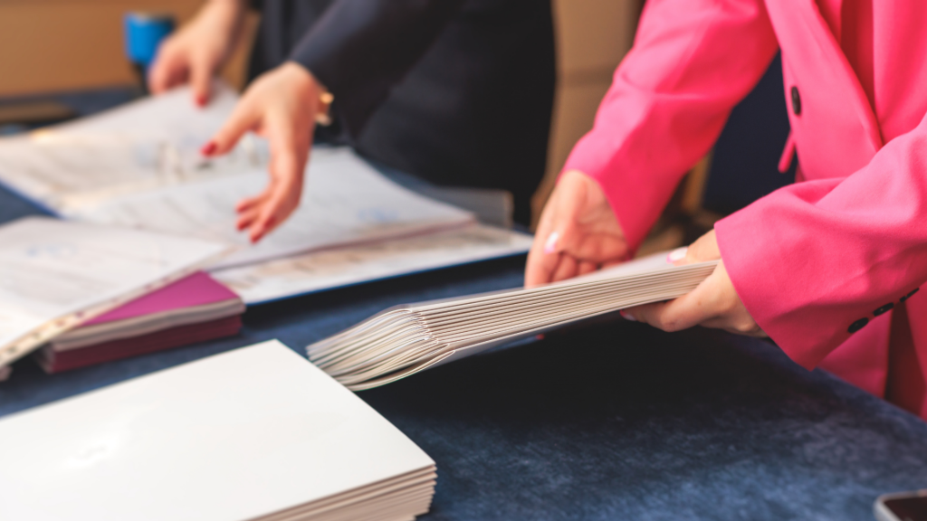Close-up of two professionals handling paperwork at a formal event. One person in a pink blazer is organizing a stack of documents, while another in dark attire is reviewing files on a table. The scene suggests business, legal, or administrative work in a professional setting.