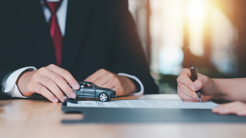 A person signing a vehicle registration document while another individual in a suit holds a small model car, symbolizing the process of vehicle registration and ownership transfer. Dirt Legal can help you register your vehicle quickly and hassle-free, making it street-legal without the DMV headaches.