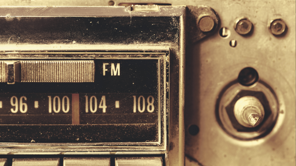 A close-up of a vintage car radio with an FM tuner, displaying frequencies between 96 and 108. The worn metal surface and aged buttons add a nostalgic feel.