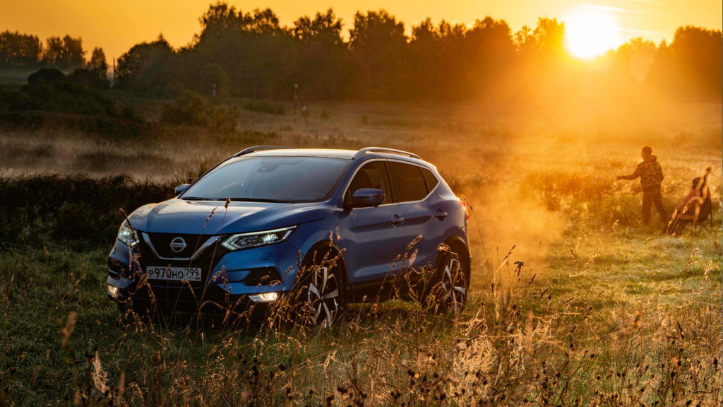A blue Nissan SUV parked in a sunlit field at sunset, surrounded by tall grass. In the background, two people walk through the dust, adding a sense of adventure and exploration to the scenic outdoor setting.