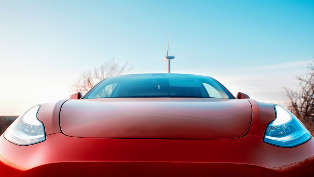 A close-up front view of a red Tesla Model 3 with its sleek headlights illuminated, parked outdoors under a clear blue sky. A wind turbine stands in the background, symbolizing clean energy and sustainability. CSS