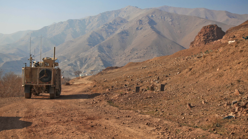 A military armored vehicle drives along a rugged dirt road in a remote mountainous region. The terrain is dry and rocky, with towering peaks in the background under a clear sky.