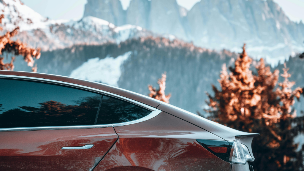 A sleek red Tesla electric vehicle is parked against a breathtaking mountain backdrop with snow-capped peaks and autumn-colored trees. The modern design of the car contrasts with the natural beauty of the landscape, symbolizing the blend of innovation and sustainability in scenic travel.