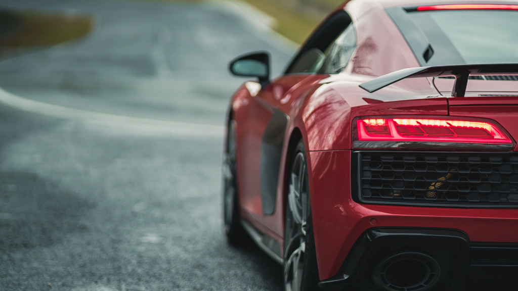 A red luxury sports car with sleek aerodynamic design and illuminated taillights is captured from the rear on a winding, wet road. The high-performance vehicle exudes speed and precision, ready for an exhilarating drive through the curves ahead.