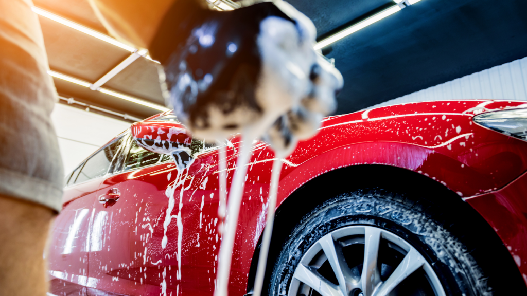 A close-up shot of a person washing a red car, with soap suds dripping from a sponge. The image captures the shine of the car’s surface, emphasizing cleanliness and detailing.