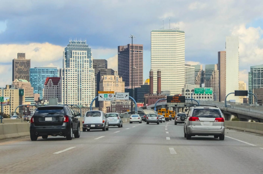 Cars driving on a busy highway leading into a city skyline with modern skyscrapers and historic buildings. The road signs and traffic indicate a metropolitan setting.