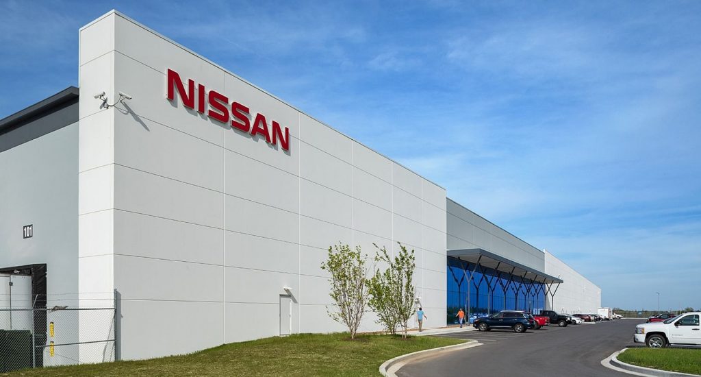 A modern Nissan manufacturing or distribution facility with a white exterior, large red Nissan logo, and a parking lot with several vehicles under a clear blue sky