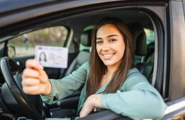 A smiling young woman sits in the driver’s seat of a car, holding up her driver's license toward the camera.