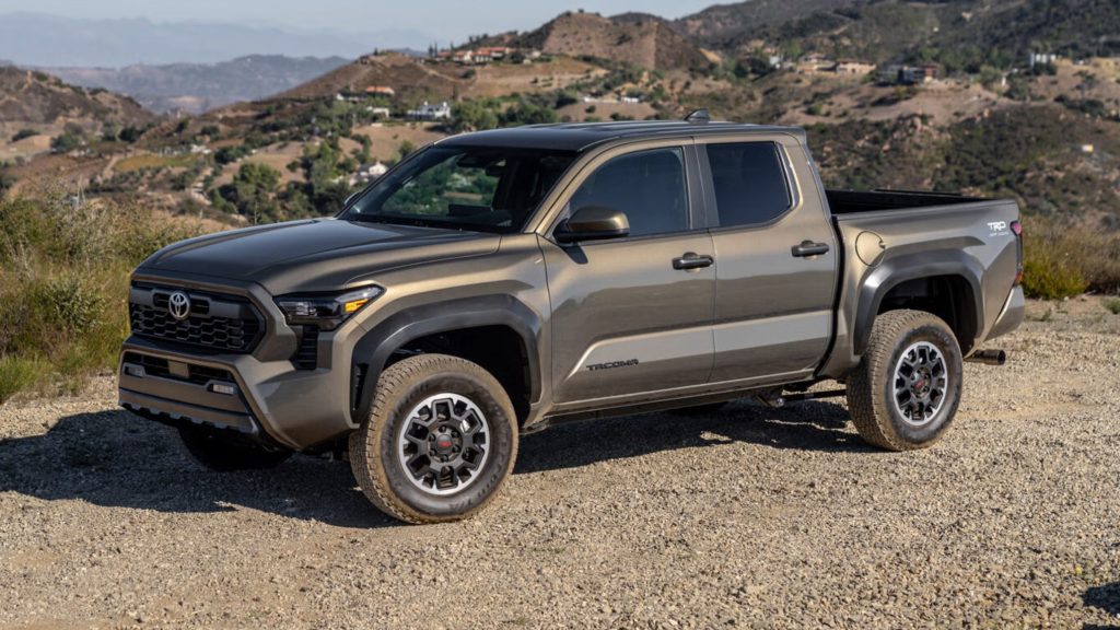 A metallic gray Toyota Tacoma pickup truck parked on a gravel overlook, with rolling hills and a scenic mountainous landscape in the background.