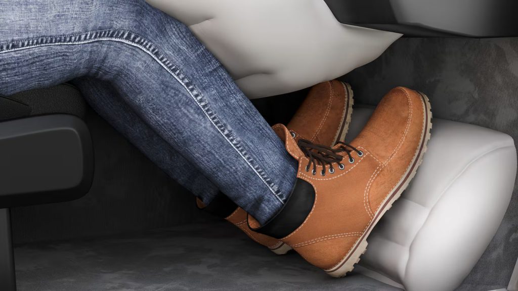 A close-up view of a person's legs wearing blue jeans and brown boots, with an activated car knee airbag deployed under the dashboard for protection.