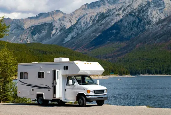 A white Class C motorhome is parked by a serene mountain lake, surrounded by lush forests and towering rocky peaks. The peaceful setting showcases the perfect backdrop for an outdoor adventure, with a boat visible in the distance on the water.