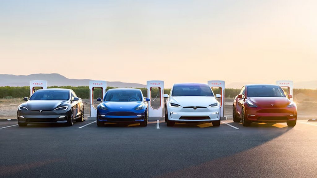 A lineup of four Tesla electric vehicles—Model S, Model 3, Model X, and Model Y—parked at a Tesla Supercharger station under a golden sunset. The EVs are plugged in, charging against a scenic backdrop of open land and distant hills, symbolizing the future of sustainable transportation.