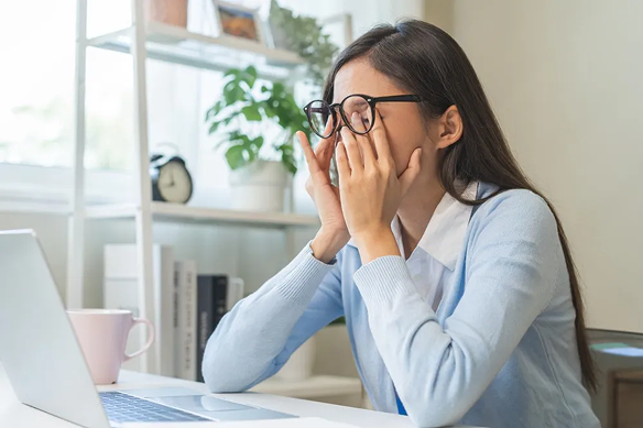 This image shows a woman wearing glasses sitting at a desk in front of a laptop rubbing her eyes in frustration or fatigue in a well-lit home office setting.