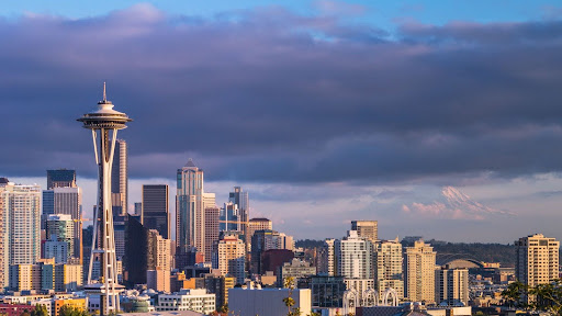 Seattle skyline featuring the Space Needle with a backdrop of modern high-rise buildings and Mount Rainier partially visible in the distance.