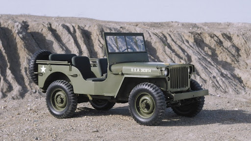 A classic military-style Willys Jeep painted in olive green, featuring an open-top design, rugged tires, and a star insignia, parked on rocky terrain with a backdrop of eroded cliffs