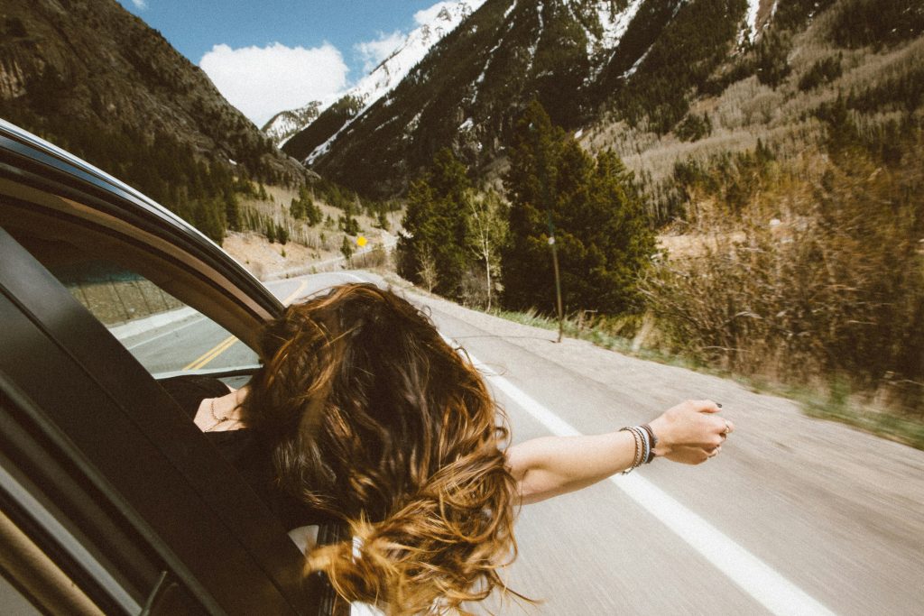 A person leans out of a moving car with their arm extended on a winding mountain road surrounded by trees and snow-capped peaks.