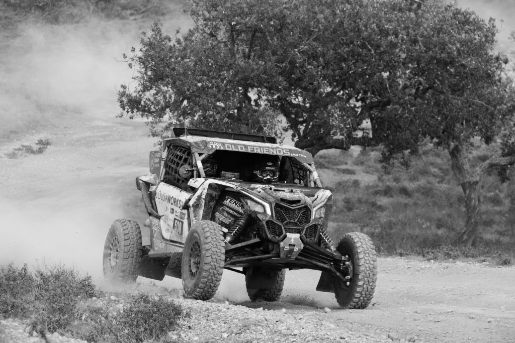 A rugged off-road side-by-side vehicle kicks up dust as it races down a dirt trail, captured in black and white with trees and rough terrain in the background.