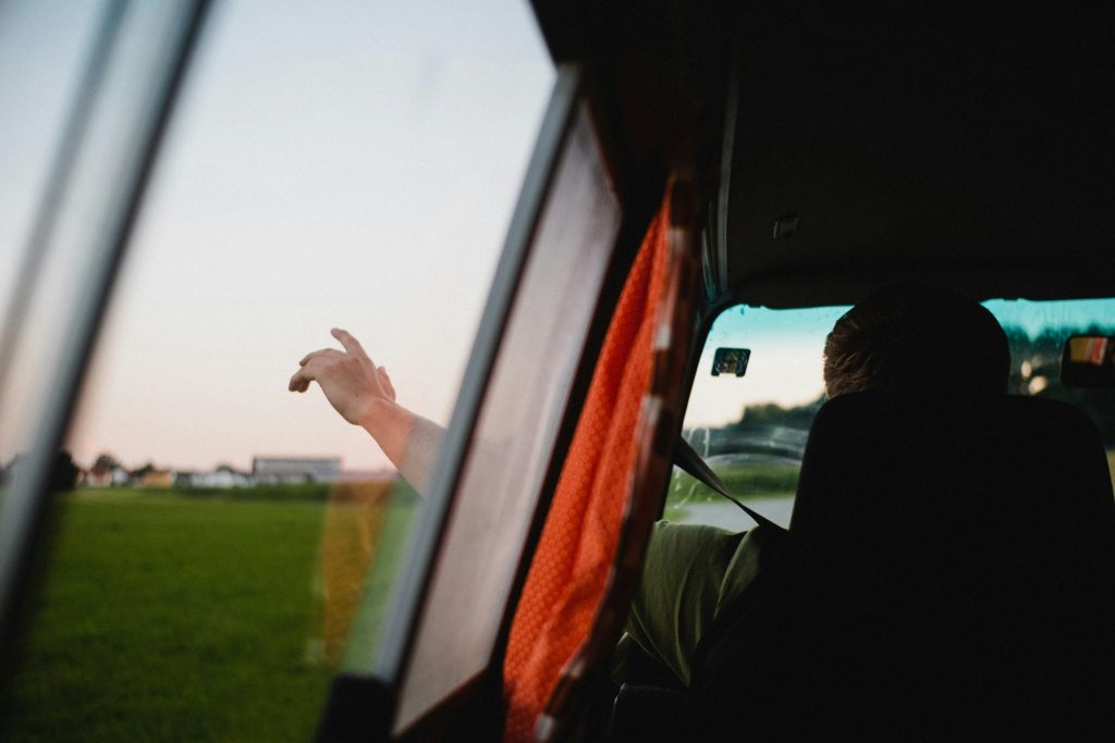 A person sits inside a vehicle with an orange curtain partially drawn while extending their arm out of the window over a green field under a soft evening sky.