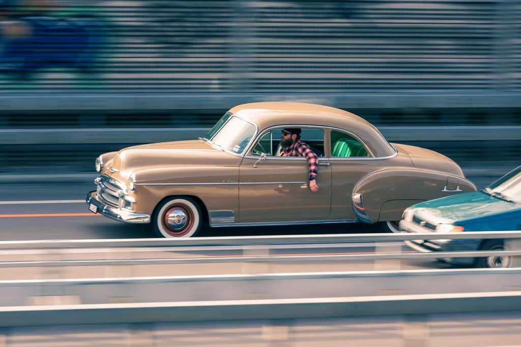A man drives a vintage beige coupe down the highway with his arm out the window.