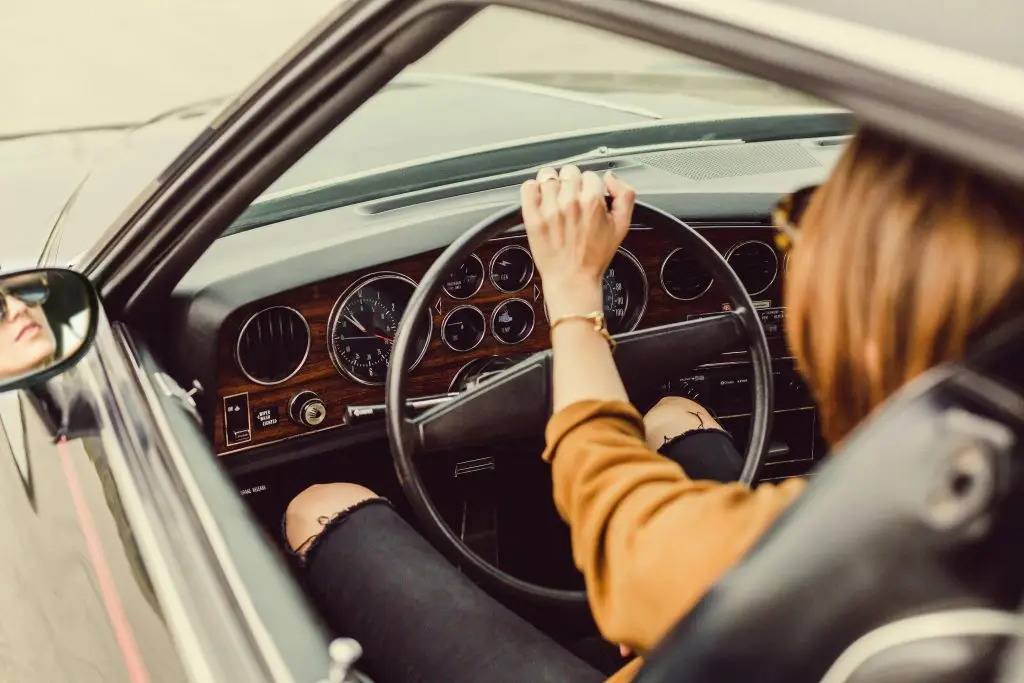 A woman drives a vintage car with a wood-paneled dashboard and classic analog gauges.