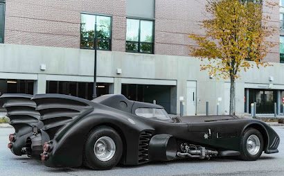 A sleek black Batmobile-style custom car with dramatic fins and an elongated body is parked on a city street near a modern brick building and a tree with autumn leaves.