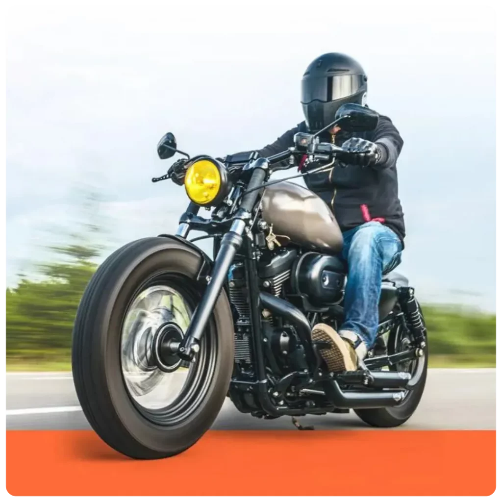 This image shows a motorcyclist wearing a black helmet and jacket riding a custom bobber-style motorcycle with a matte tank and yellow headlight on an open road.