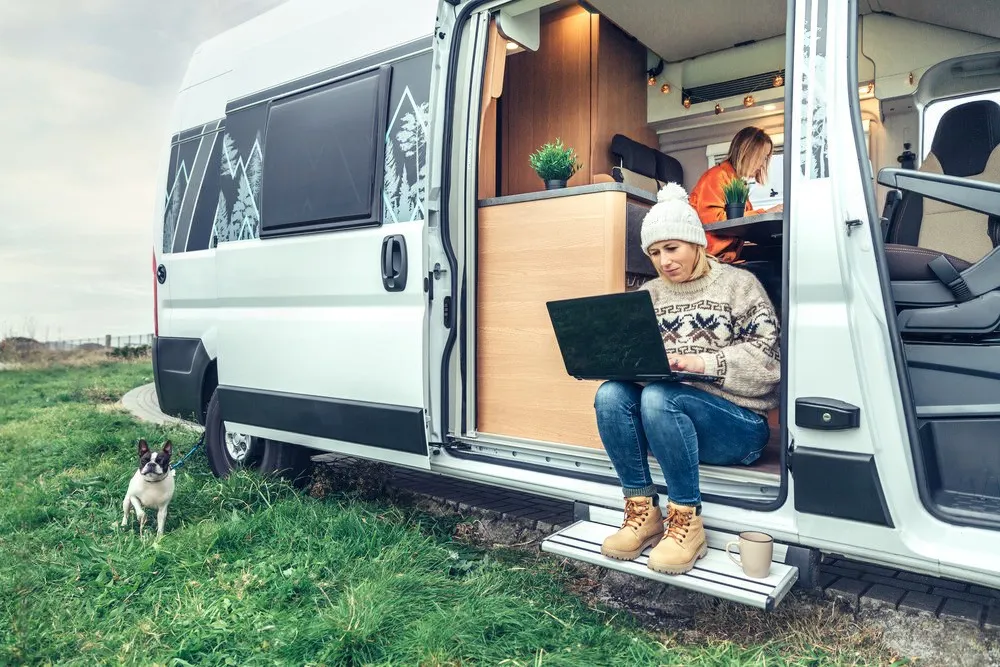 This image shows a woman working on a laptop while sitting on the step of a camper van, with another person inside working at a table and a small dog on a leash nearby, capturing a cozy and modern van life scene in a grassy outdoor setting.