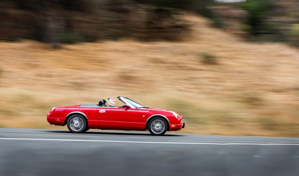 A red convertible sports car speeds along a highway with its top down, captured in motion against a blurred, dry hillside backdrop.