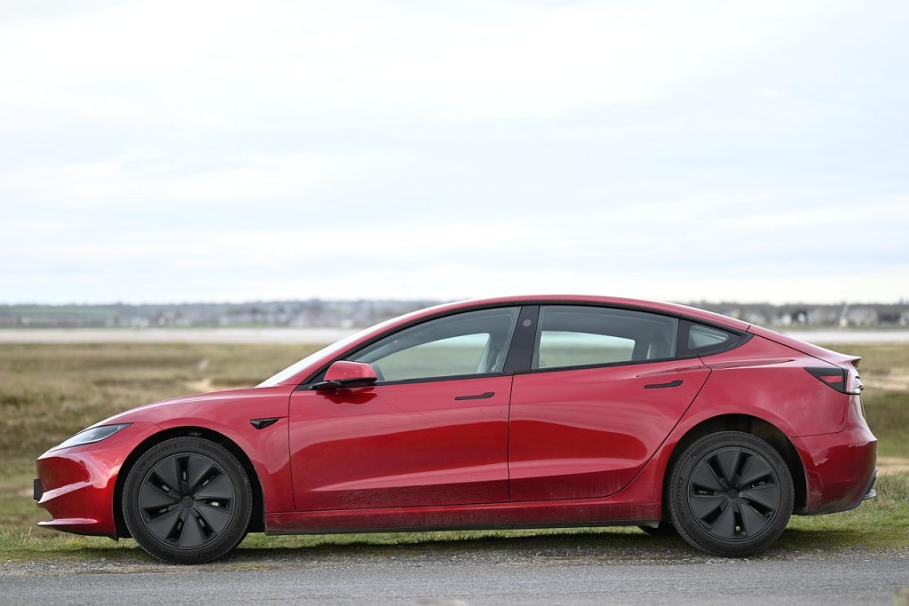 A red Tesla Model 3 is parked on a rural roadside with open fields and a distant town visible in the background.