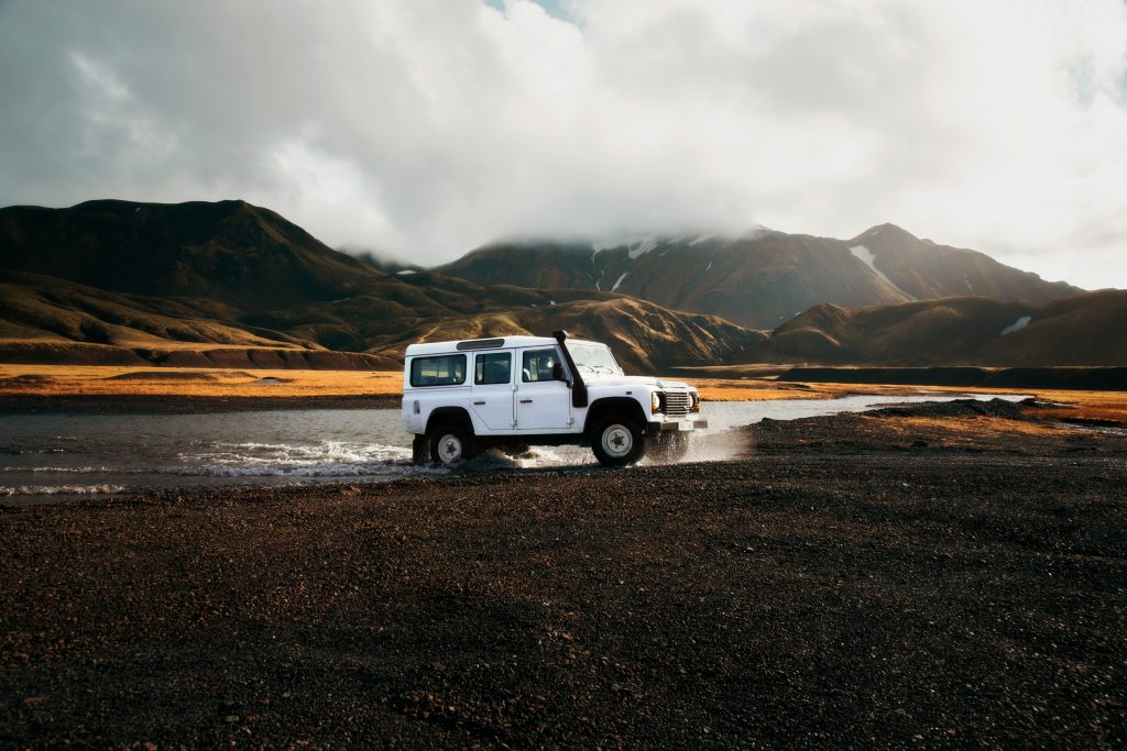 A white off-road SUV drives through a shallow stream in a remote, rugged landscape with dramatic mountains and moody clouds in the background.