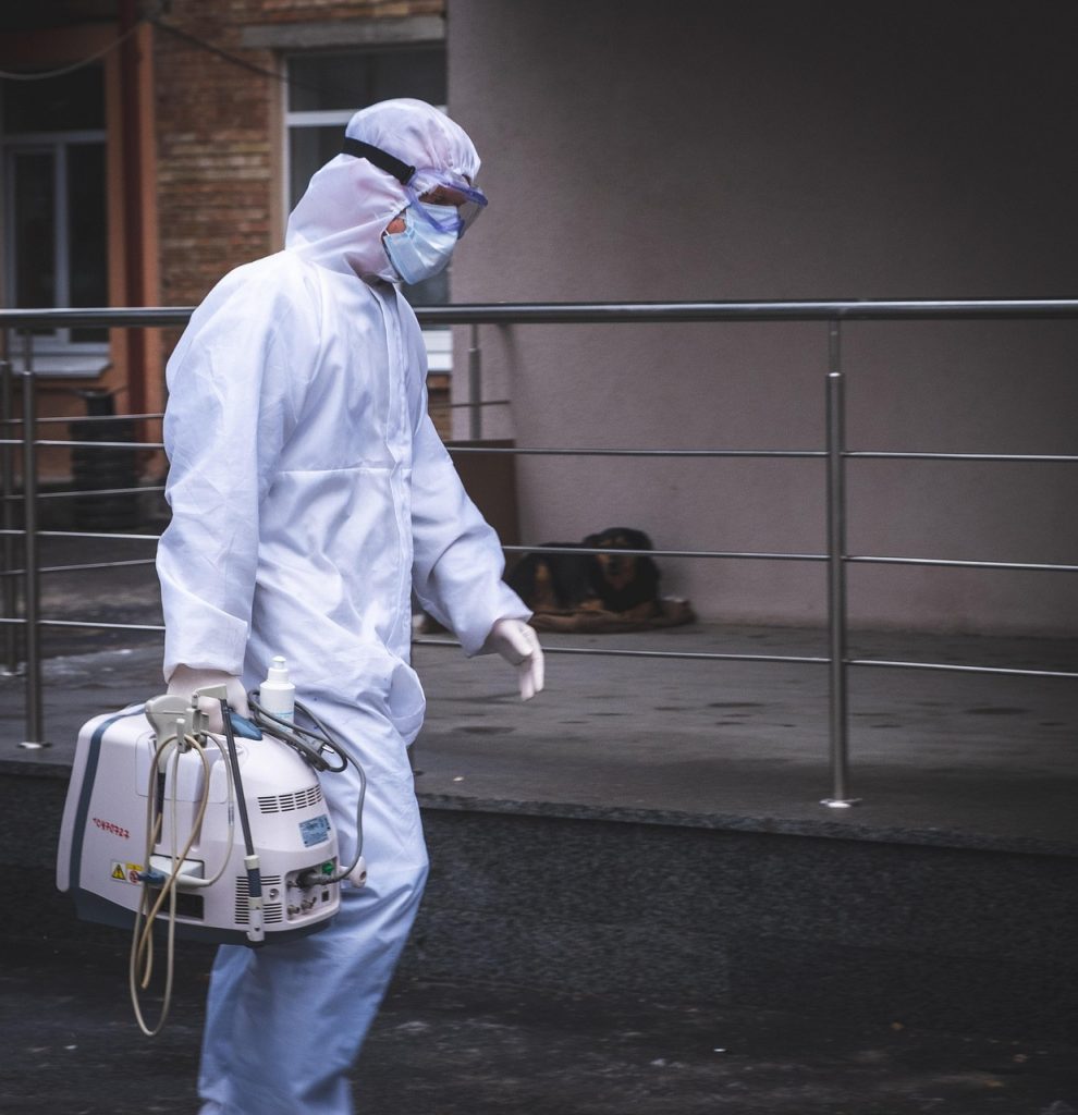 A healthcare worker in full protective gear carries a portable medical device outside a building, emphasizing caution and preparedness.