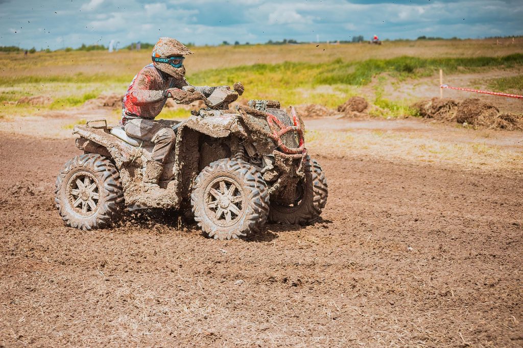 A mud-covered ATV rider speeds through a rugged off-road track, surrounded by open fields under a cloudy sky.