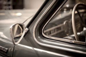 This image shows a close-up of a vintage car’s driver-side window, side mirror, and steering wheel, highlighting its classic chrome trim and retro interior design.