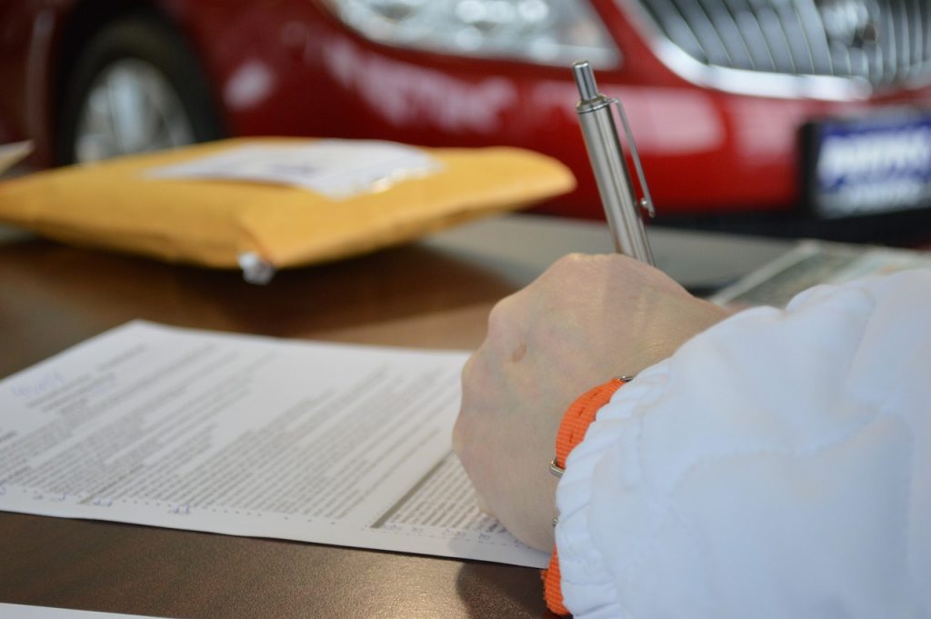A person signs paperwork at a desk with a red car and mailing envelope in the background, suggesting a vehicle purchase or registration process.