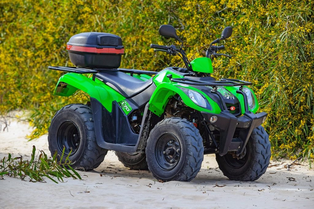 A bright green ATV with rugged tires and a rear cargo box is parked on sandy terrain, ready for off-road adventure near a dense backdrop of yellow-flowered bushes.