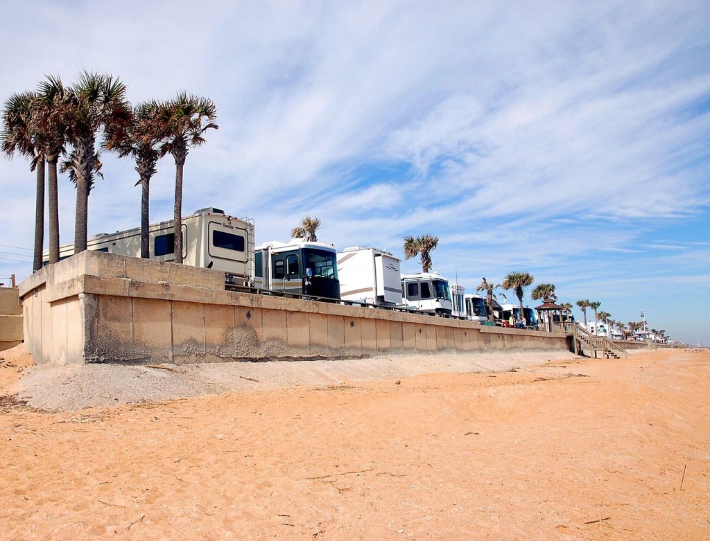 A line of RVs is parked along a beachfront wall under a bright blue sky, with palm trees swaying nearby and the sandy shore stretching out below.