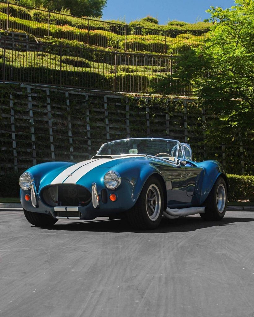 This is a blue Shelby Cobra replica with white racing stripes, parked on a sunny day in front of a green hillside.