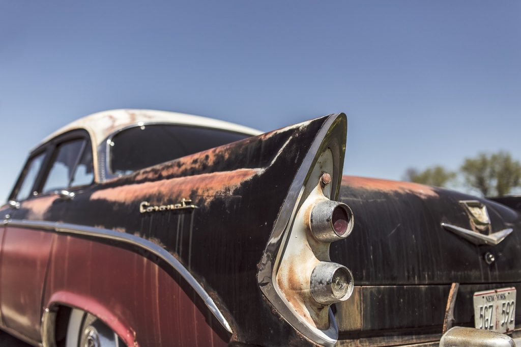 A weathered classic car with faded red and black paint and rusted chrome tailfins sits under a clear blue sky, evoking vintage charm and untold stories from the road.