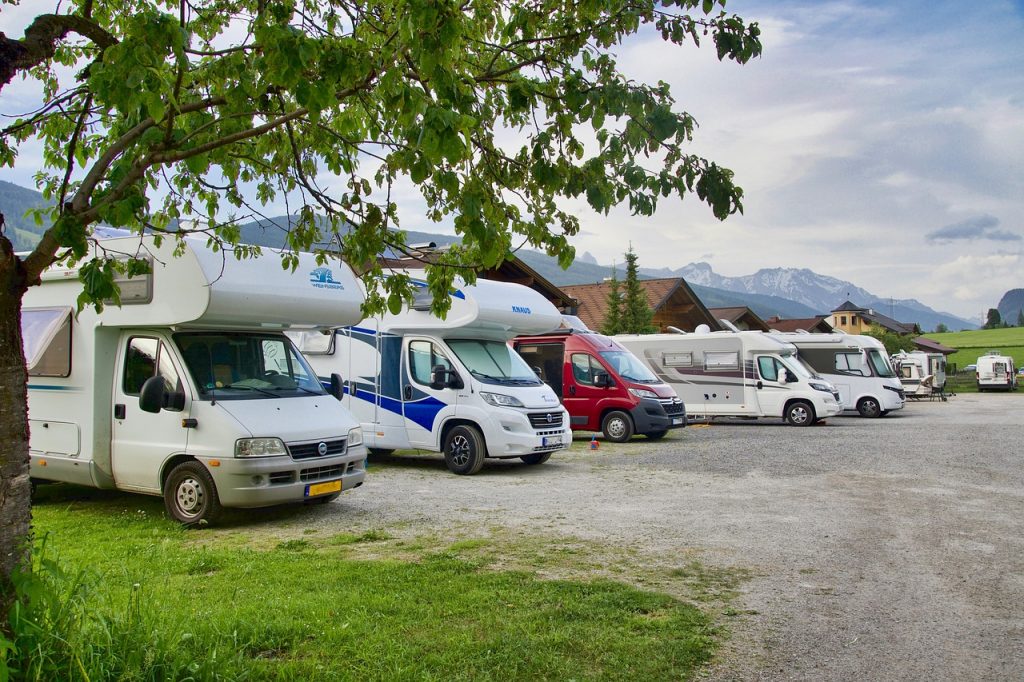 A row of RVs is parked in a scenic lot surrounded by mountains, trees, and quaint wooden houses, offering a peaceful camping atmosphere.