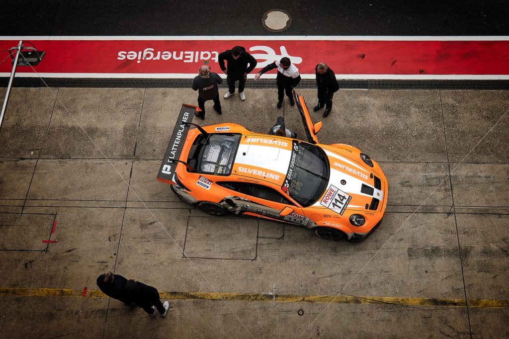 An overhead view of an orange race car in the pit lane shows a small crew gathered around preparing it for the track.