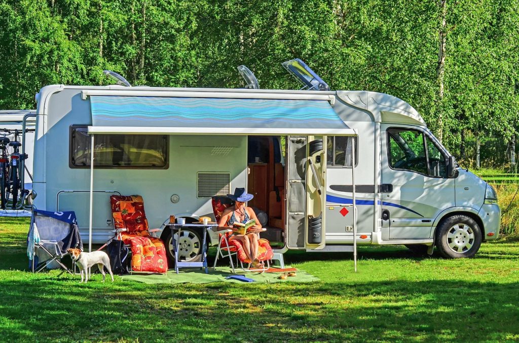 A person relaxes under the extended awning of their RV, enjoying the sunshine with outdoor chairs, a table, and a dog nearby in a peaceful forested campground.