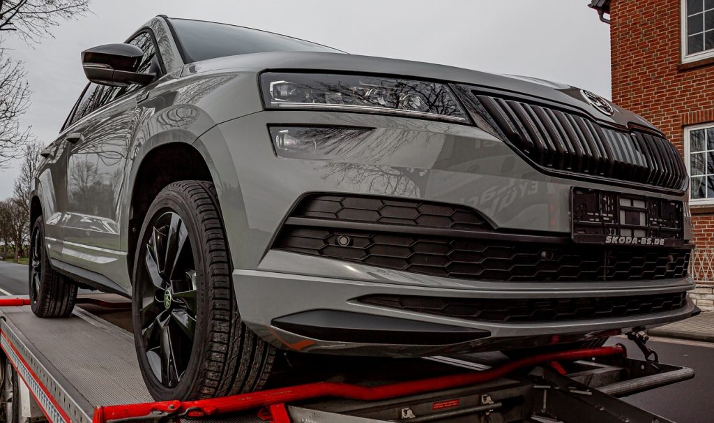 A sleek gray SUV with black accents is secured on a red flatbed trailer, likely in transit for delivery or registration, parked near a brick building under an overcast sky.