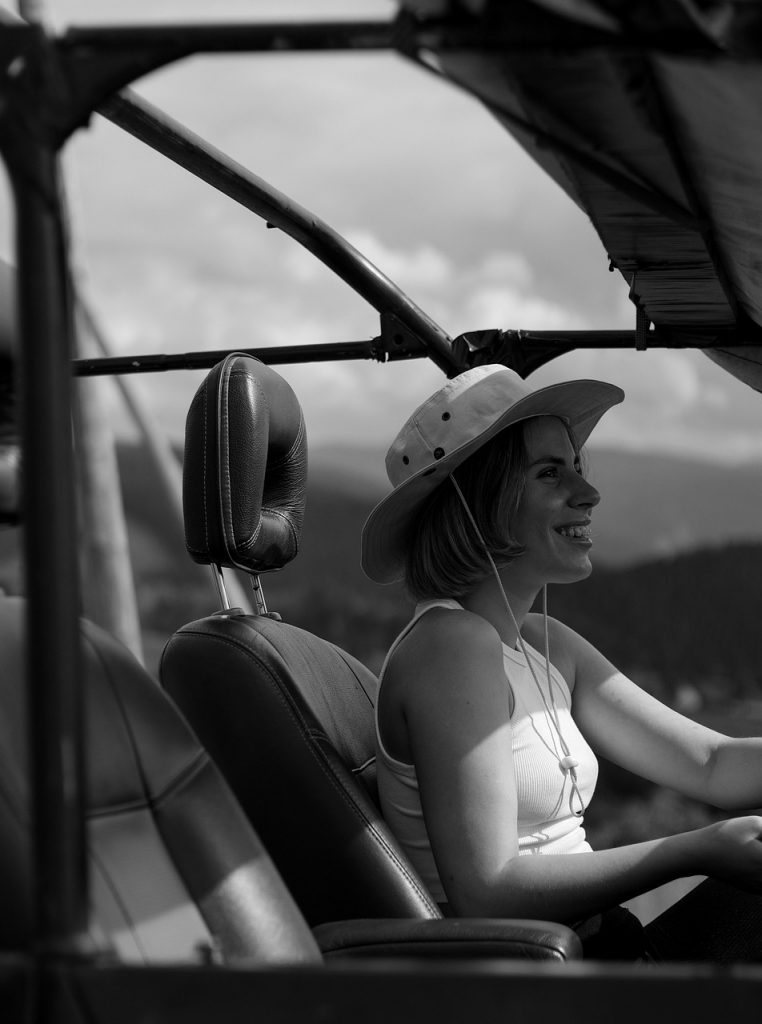 A woman wearing a wide-brimmed hat smiles as she drives an open-air off-road vehicle through a scenic mountain landscape.