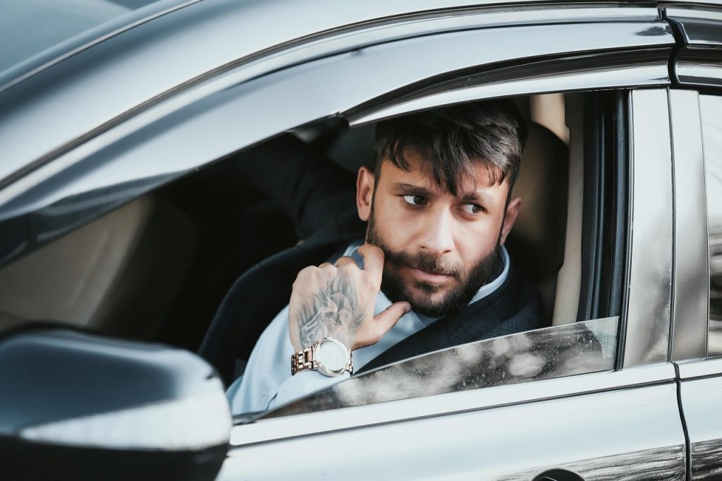 A confident man with tattoos and a stylish watch sits in a sleek black car, gazing out the window with determination—ready to hit the road with registration handled.