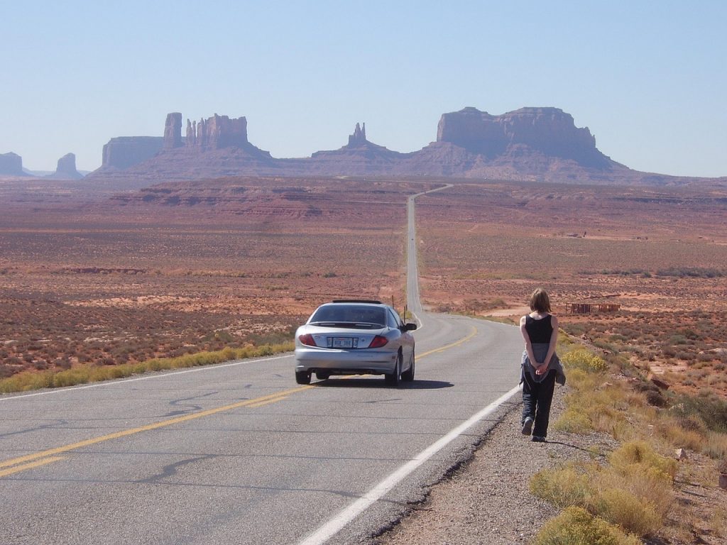 A silver car and a lone traveler face the open desert road toward Monument Valley’s towering red rock formations, evoking the freedom and savings possible with Montana LLC registration.